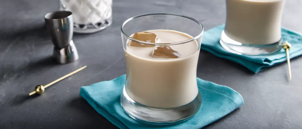 Two glasses of White Russian with ice on a blue napkin, accompanied by cocktail picks and a jigger