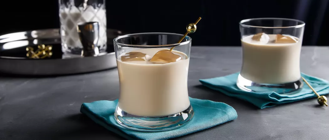 Two glasses of White Russian with ice, both on a blue napkin. One is with a cocktail pick, and the other is alongside a cocktail pick