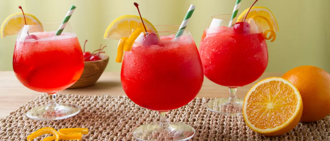Three snifter glasses of Sunset Daiquiri placed on a jute mat, garnished with cherry, lemon slice, orange peel with a straw. Alongside an orange, halved orange with some peeled orange and a wooden bowl filled with cherries.