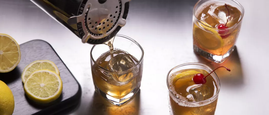 person pouring Amaretto Sour into glass alongside two glasses of Amaretto Sour and a wooed board with sliced lemon on it