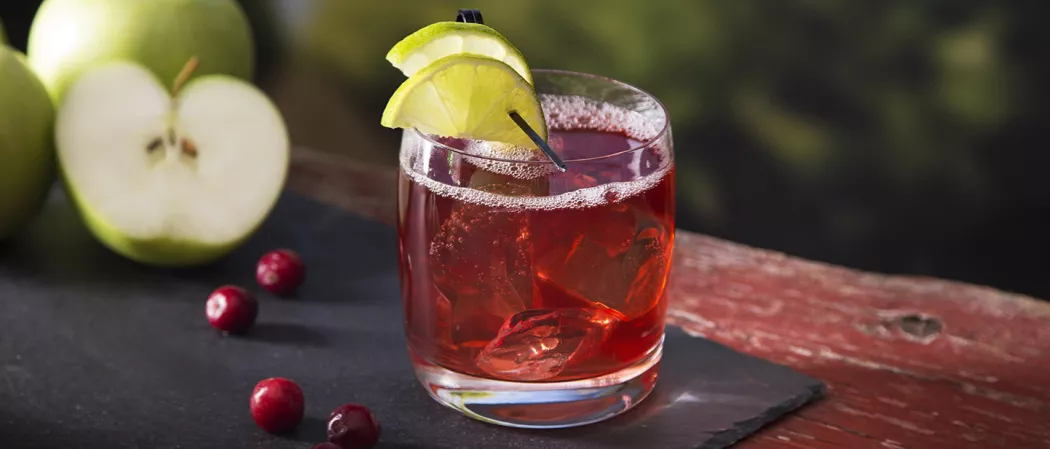 A glass of Orchard Twist garnished with lemon slices, containing crushed ice and accompanied by a few cranberries and green apples on the black tray.