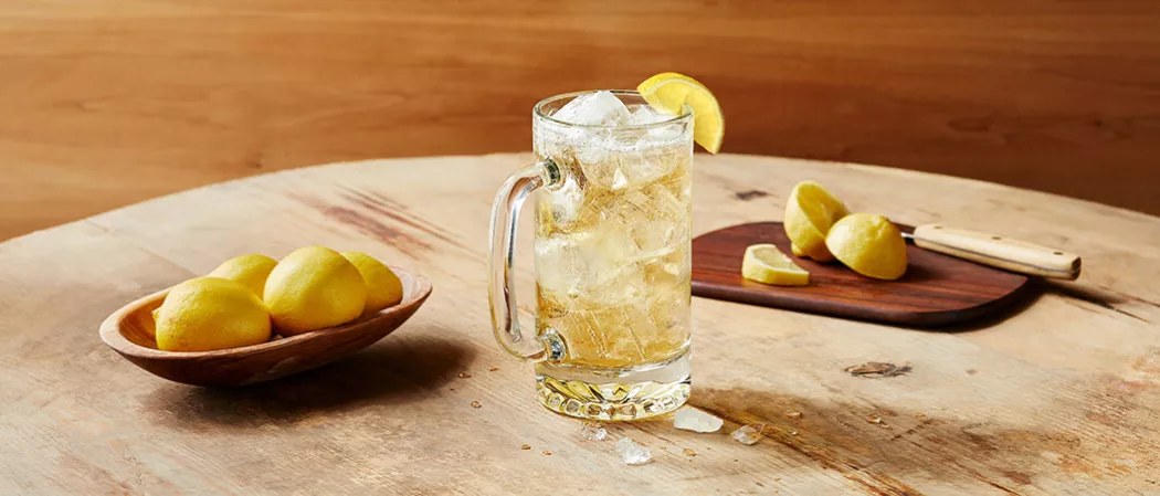 A glass of Jim Beam® and Ginger Highball garnished with lemon slice alongside wooden bowl of lemons and a chopping board with knife and chopped lemons