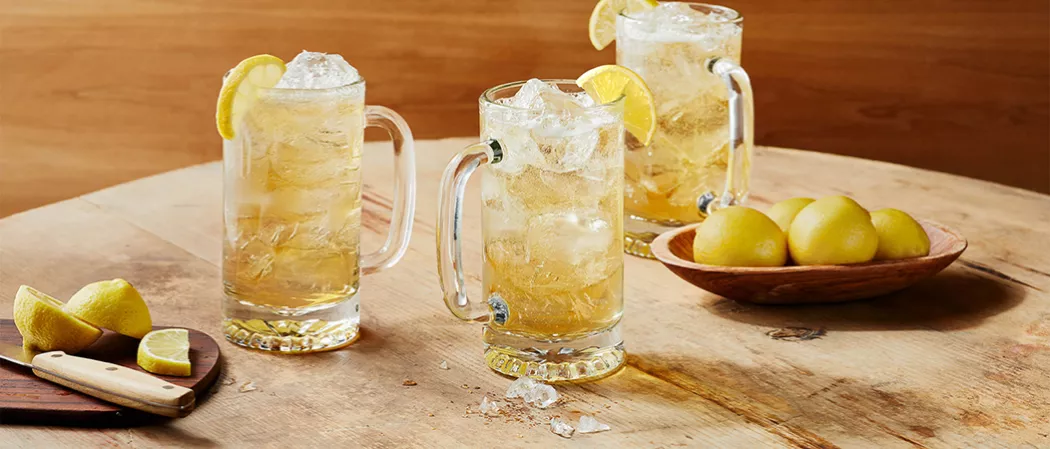 Three glasses of Jim Beam® and Ginger Highball garnished with lemon slice alongside wooden bowl of lemons and a chopping board with knife and chopped lemons