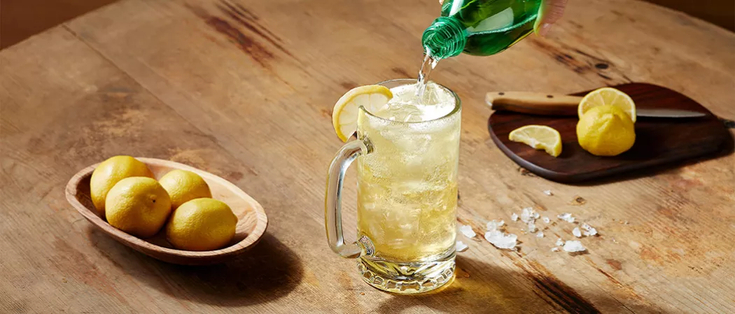 A person pouring Ginger Ale into the glass of Jim Beam® and Ginger Highball  alongside wooden bowl of lemons and a chopping board with knife and chopped lemons