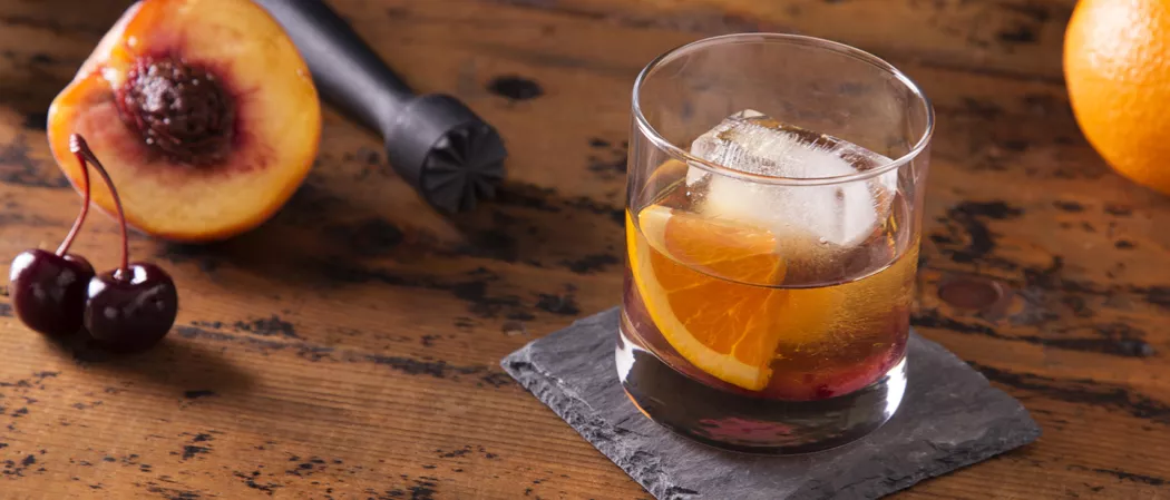 A glass of Peach Old Fashioned garnished with orange wedge on rock coaster alongside half peach and two cherries