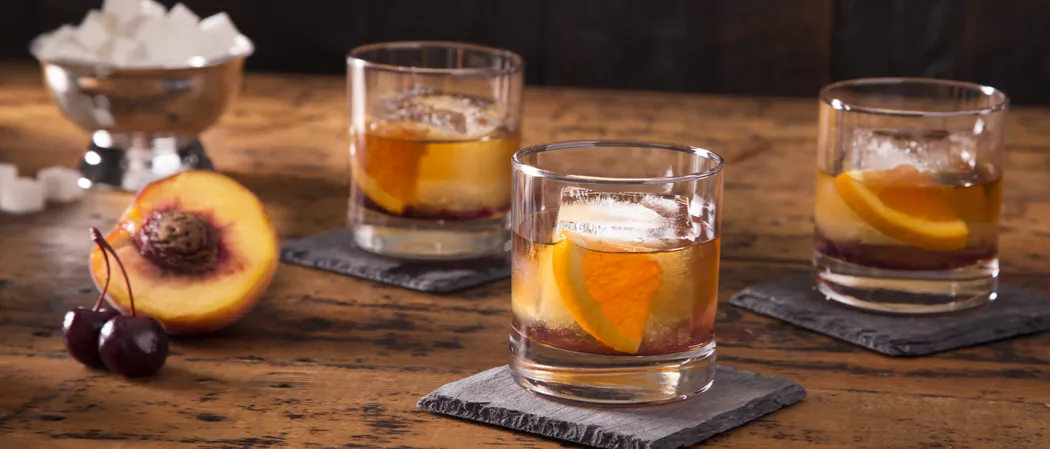 Three glasses of Peach Old Fashioned garnished with orange wedge on rock coaster alongside half peach and two cherries
