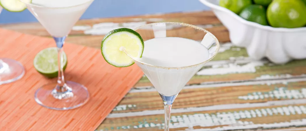 Two glasses of Key Lime Pie Martini, each garnished with whipped cream and a lime wheel on the rim of the glass, alongside slice of lime and a  bowl full of lime