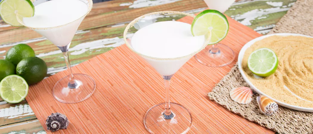 Two Key Lime Pie Martinis, garnished with whipped cream and a lime wheel, placed with lime on graham crackers and shells. Lime wedges and slices arranged on table mat.