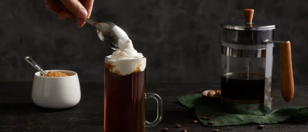 A person garnishing Whipped heavy cream on the top of Kilbeggan® Irish Coffee along with Brown Sugar Syrup