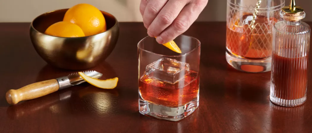 A person adding lemon peel into glass of Knob Creek® Old Fashioned alongside a peeler, a bowl of lemons.