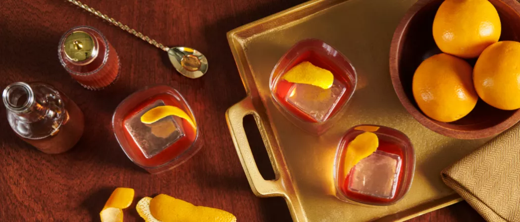 Three glasses of Knob Creek® Old Fashioned garnished with lemon peel and ice, two placed on on a golden tray with a bowl of lemons. alongside a glass of same drink, a bar spoon, and few lemon peels