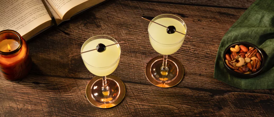 Two glasses of Last Word garnished with black cherries along with the small dry fruits bowl on a green cloth , and a candle jar