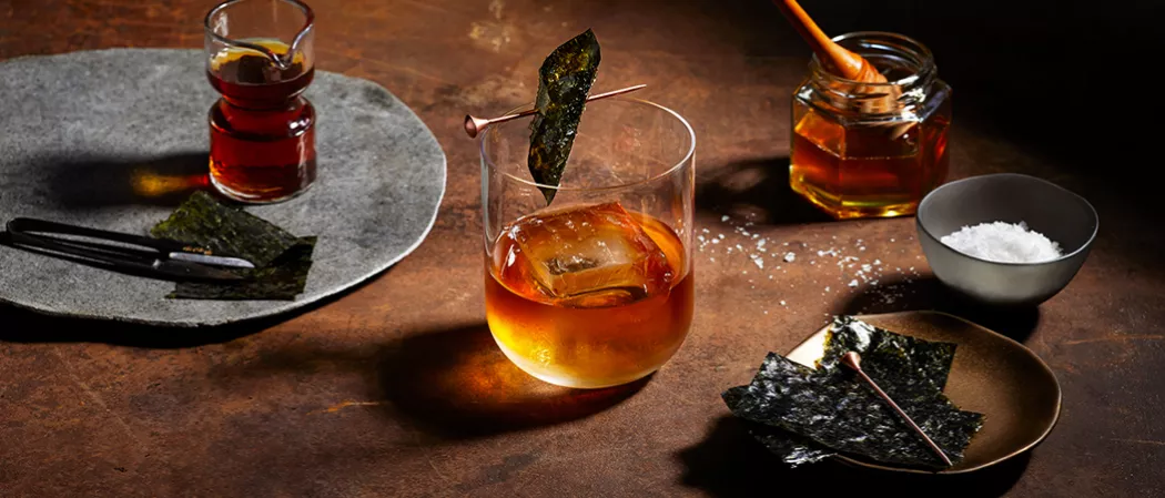 A glass of Legent® Sea 'New' Fashioned garnished with Nori Strip along with Orange Bitter and a small  bottle of Salted Honey Syrup  and a bowl of Nori Strips