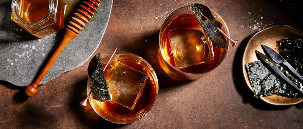 Two glasses of Legent® Sea 'New' Fashioned garnished with Nori Strip along with bottle of Salted Honey Syrup  and a bowl of Nori Strip 