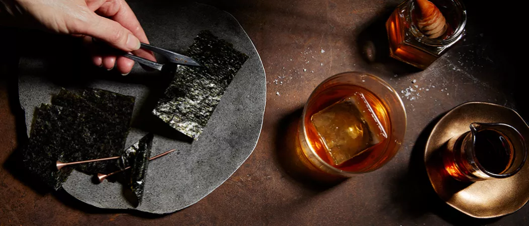 A glass of Legent® Sea 'New' Fashioned garnished with Nori Strip along with Orange Bitter and a small  bottle of Salted Honey Syrup  and a bowl of Nori Strip 
