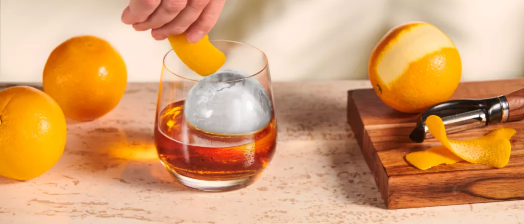 A person adds orange peel to a Legent® Blossom Old Fashioned glass with two oranges, one peeled, on a wooden board.