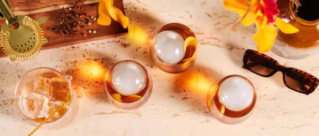 Three glasses of Legent® Blossom Old Fashioned on a wooden board with a sunglass, a strainer, various spices, orange peel  and another glass of ice cubes with a golden stirrer