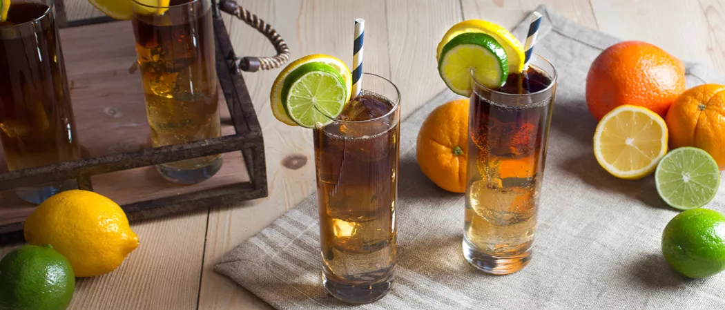 Two glasses of Long Island Iced Tea with a straw, garnished with a lime and lemon wheel on a mat, alongside few lime, lemon, and orange slices. Accompanied by two glasses on a tray.