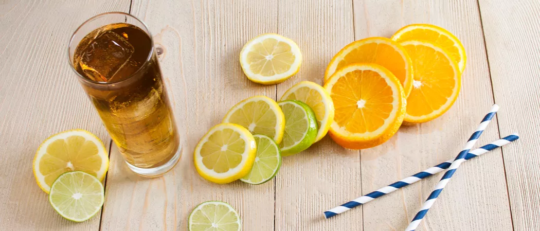 Glasses of Long Island Iced Tea with ice cubes, alongside some lemon, lime, and orange wheels, and two straws.