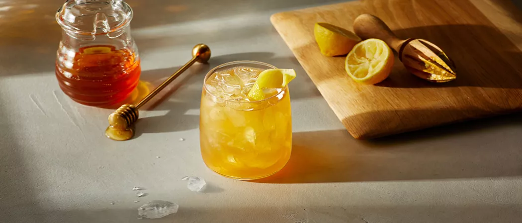 Glass of Maker's Mark® Gold Rush garnished with lemon peel alongside honey pot, honey dipper and wooden board with lemon and hand juicer on it.
