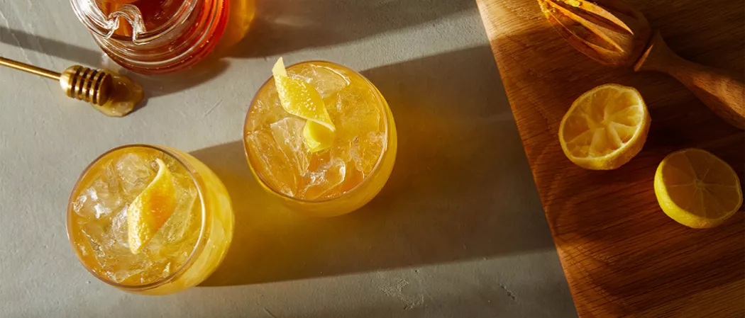Two Glasses of Maker's Mark® Gold Rush garnished with lemon peel alongside honey pot, honey dipper and wooden board with lemon and hand juicer on it
