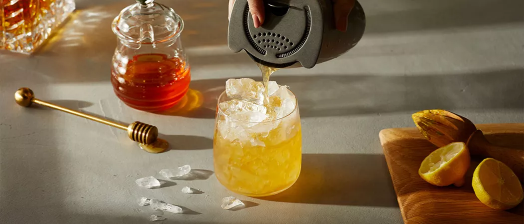 A person pouring Maker's Mark® Gold Rush into glass filled with ice alongside honey pot, honey dipper and wooden board with lemon and hand juicer on it.
