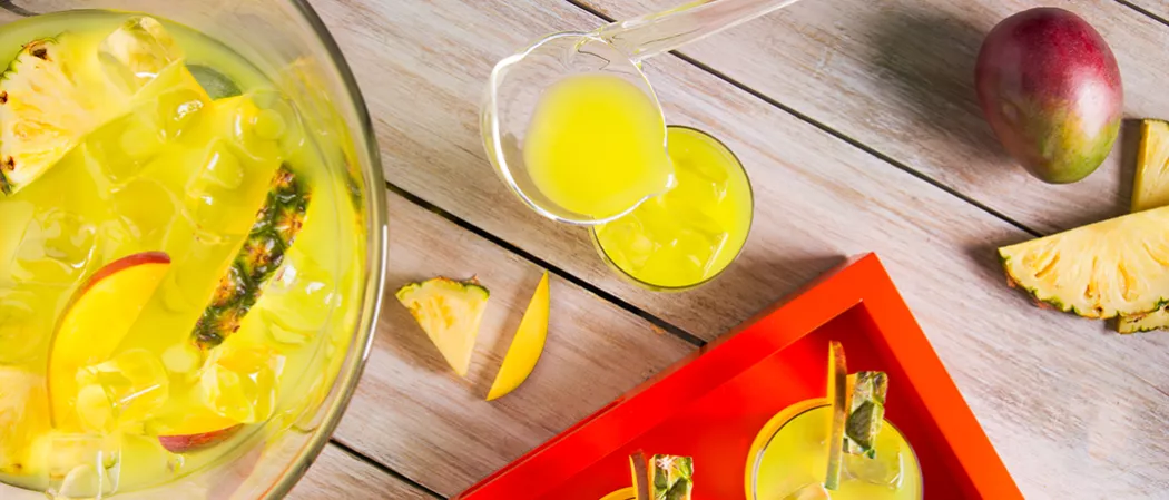 A person is pouring Pina Punch into a glass with ice, alongside a glass of Pina Punch on a red tray,  few mango and pineapple slices, and a container of Pina Punch with ice, pineapple, and mango slices.