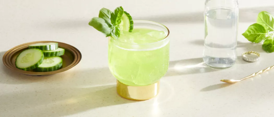 A glass of Midori® Pot of Gold garnished with cucumber and mint Basil leaves along with chopped cucumber in a copper plate