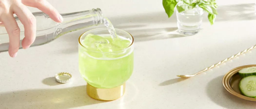 A person Pouring a soda water into a glass of Midori® Pot of Gold along with the stirring spoon on the table
