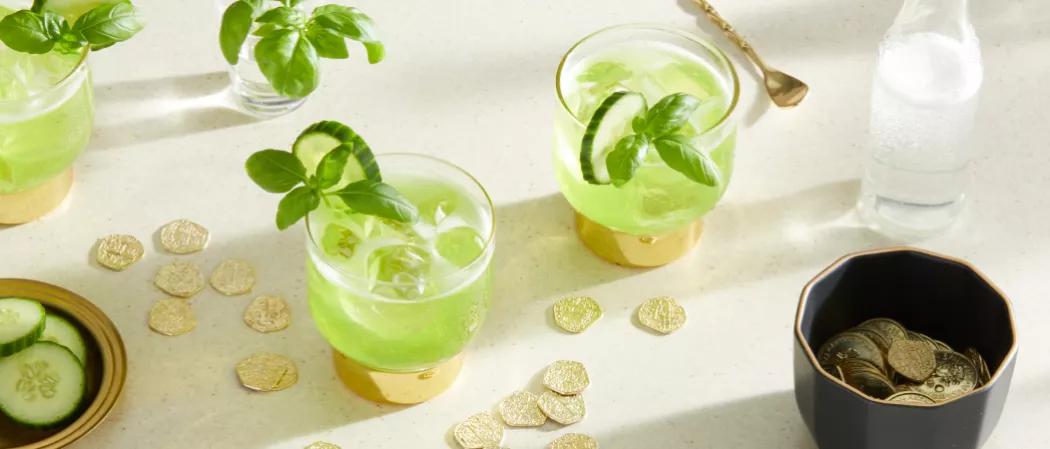 Three glasses of Midori® Pot of Gold garnished with cucumber and basil leaves along with soda bottle and a bowl of chocolates