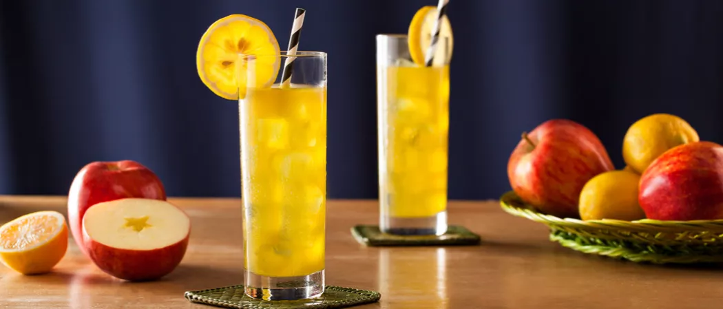 Two glasses of Midori® Cider with strawss on coasters, garnished with lemon wheels. Nearby, there is one and half apple, half a lemon and a tray of apples and lemons.