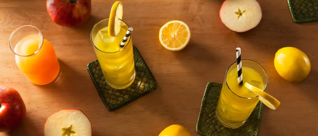 Two glasses of Midori® Cider with strawss on coasters, garnished with lemon wheels. Nearby is a jar of apple cider , few apples and some lemons 