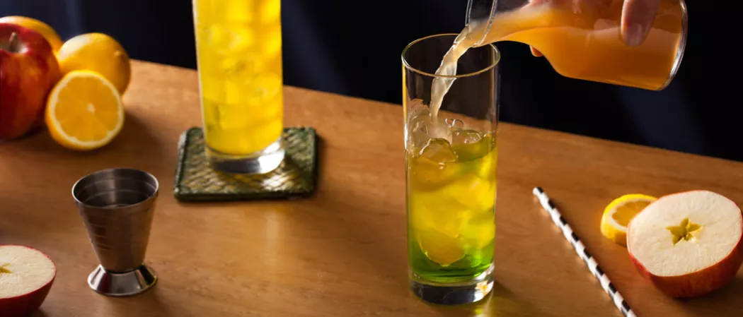 Two glasses of Midori® Cider. One glass is on a coaster, and the other contains ice being poured into with apple cider. Nearby are a jigger, a straws, and a few apples and lemons.