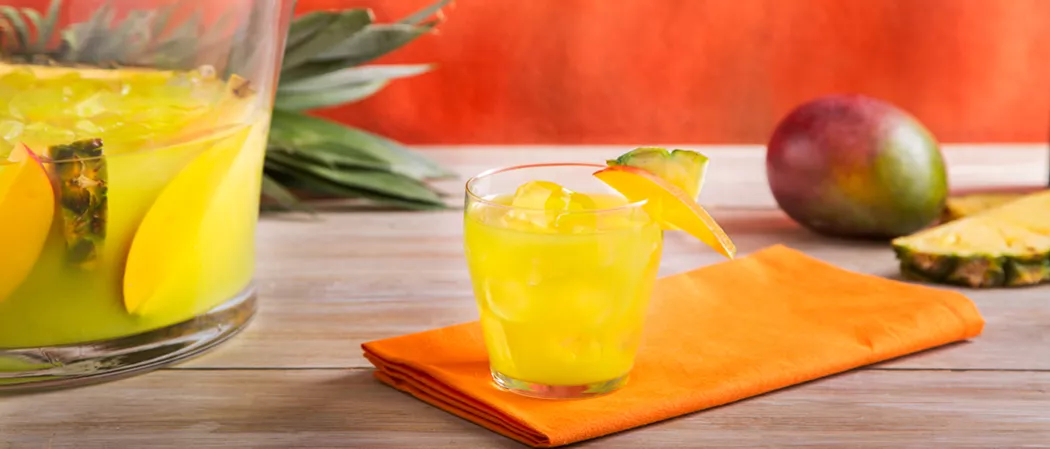 Glass of Pina Punch with ice, garnished with a mango and pineapple slice on an orange napkin. Nearby a mango,  few pineapple slices, and a container of Pina Punch with ice, pineapple and mango slices.