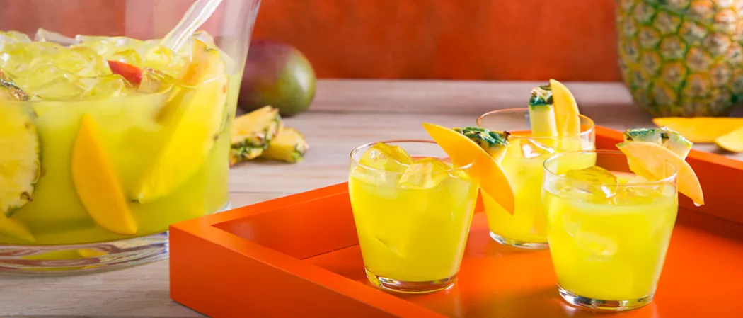 Three glasses of Pina Punch with ice, garnished with mango and pineapple slices on a red tray. Nearby a mango, few pineapple slices, and a container of Pina Punch with ice, pineapple and mango slices.