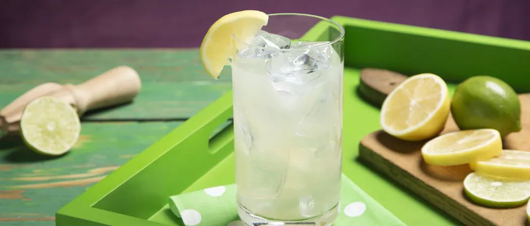 A glass of Limonlicious garnished with Lemon Wedge and there chopped lemons and a sqeezer on the table