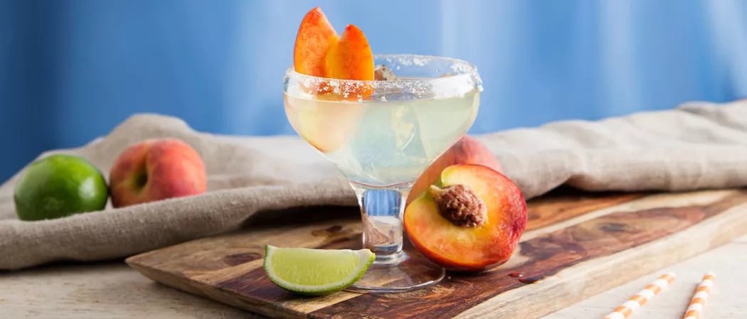 A glass of Sauza® Peach Margarita, garnished with wedges of peach, accompanied by a slice of lime and a slice of peach, placed on a wooden tray. Additionally, lemon and peach slices are arranged on a cloth.