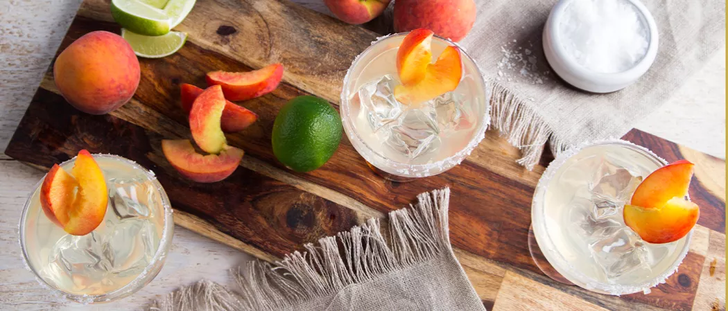 Three glasses of Sauza® Peach Margarita, each garnished with wedges of peach and ice cubes. Two glasses are placed on a wooden tray, accompanied by a peach and lime.
