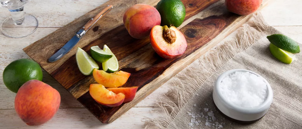 One peach and lime, accompanied by wedges of lime and peach and a knife, are all placed on a wooden tray. Additionally, a bowl of salt and a few slices of lime are placed on a cloth.
