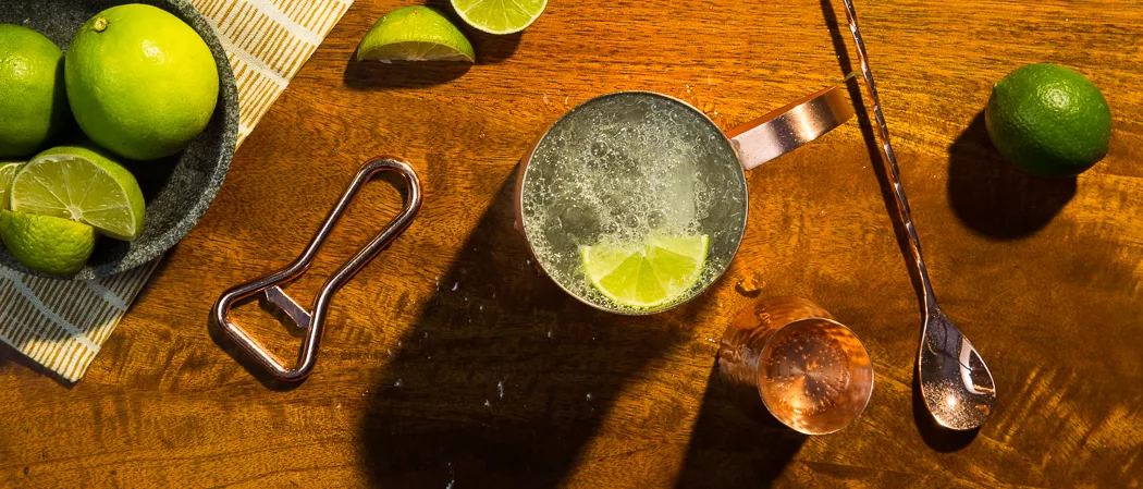 A copper cup of Pinnacle® Mule garnished with Lime Wedge along with a bowl of lemons and a stirrer with some chopped lemons