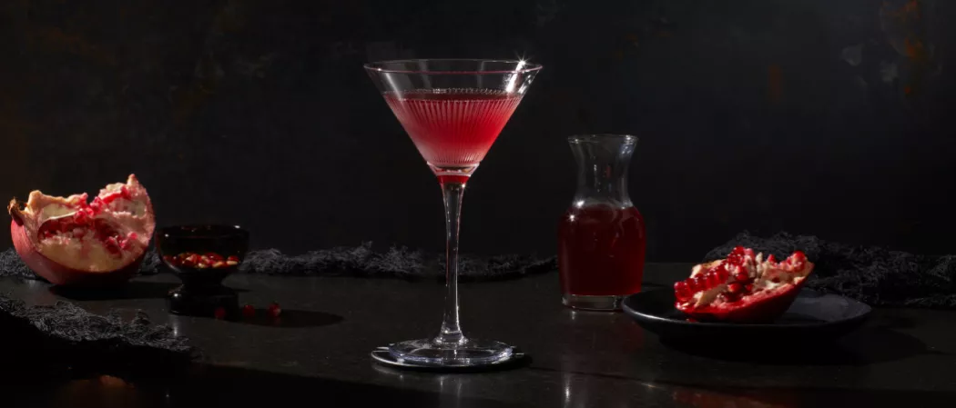  Glass of Pinnacle® Black Widow Martini alongside pomegranate in a black plate and small bottle of same drink