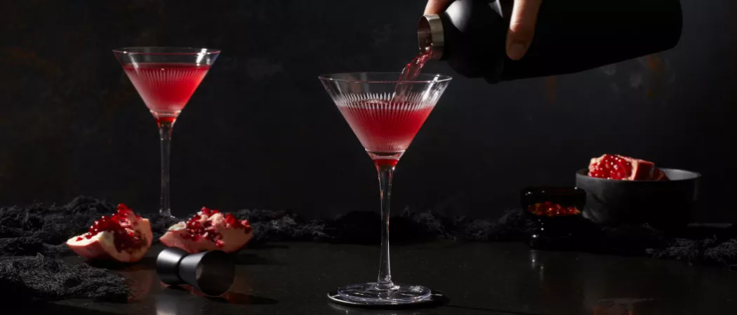 A person pouring Pinnacle® Black Widow Martini into a glass alongside another glass of Pinnacle® Black Widow Martini, few pomegranate and a jigger