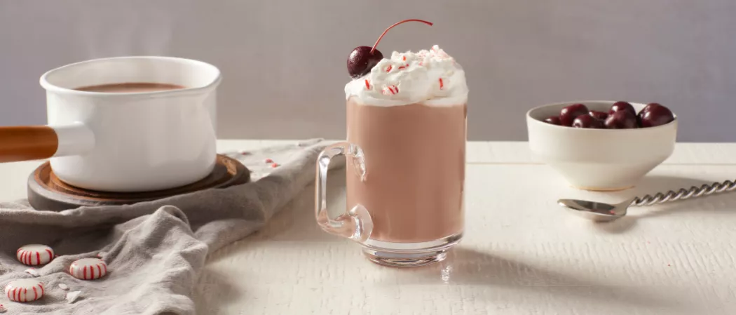 A glass of Pinnacle® Peppermint Hot Chocolate garnished with Whipped Cream and cherry along with  bowl of Swiss Miss® Milk Chocolate Hot Cocoa in a pan and bowl