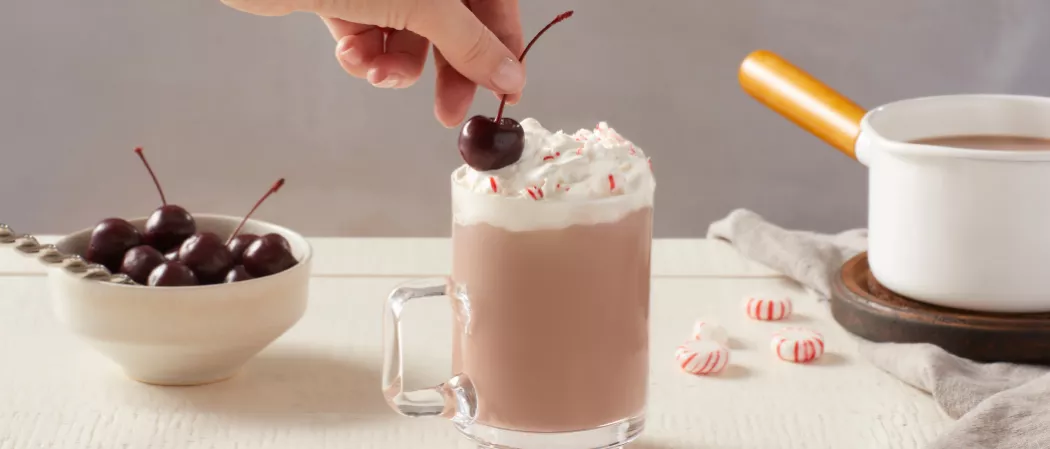 A person garnishing Cherry on a glass of Pinnacle® Peppermint Hot Chocolate along with a bucket of Cherries