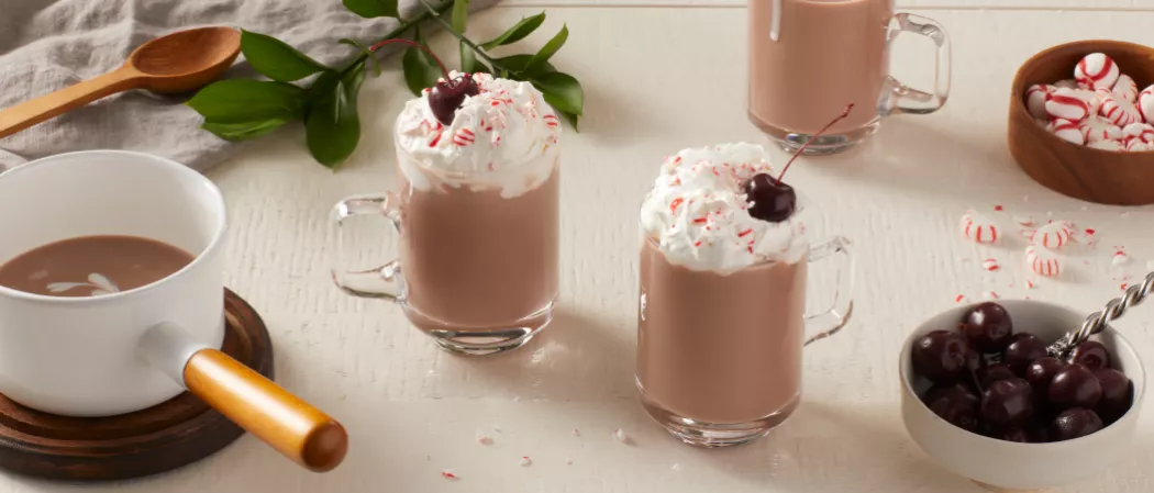 Two glasses of Pinnacle® Peppermint Hot Chocolate garnished with Whipped Cream and cherry along with  bowl of Swiss Miss® Milk Chocolate Hot Cocoa in a pan and bowl