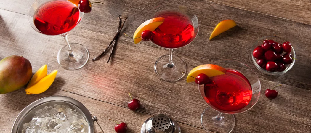 Three glasses of Mango Cran-Tini garnished with mango slice and cherry. Nearby a mango, mango slices, few cherries, cocktail sticks, a bowl of cherries, and a bucket of ice.