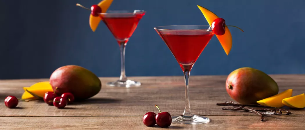 Two glasses of Mango Cran-Tini garnished with mango slice and cherry. Nearby few mangos, mango slices, few cherries and cocktail sticks.