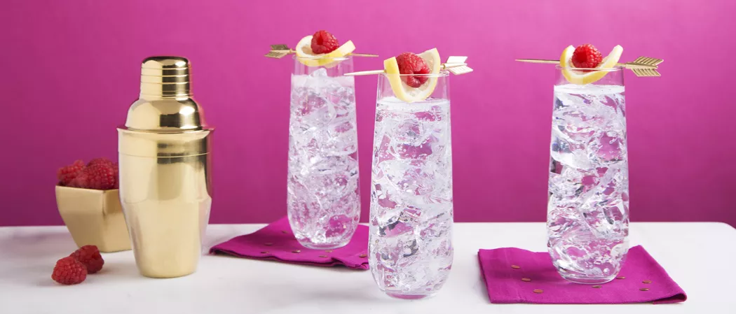 Three glasses of Citrus Fizz garnished with raspberry and lemon slice alongside bowl of raspberries and a shaker