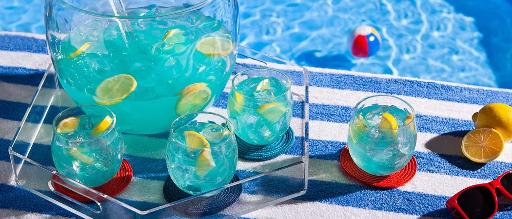 A tray by the pool holds a summer fishbowl, three glasses with ice-cold drinks and sliced lemons, and an extra glass of the same drink.