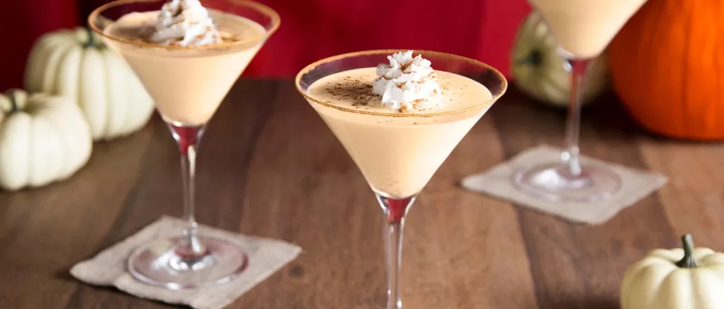 Two glasses of Pumpkin Pie Martini, each garnished with whipped cream and Pumpkin Spice, both placed on a small piece of cloth.